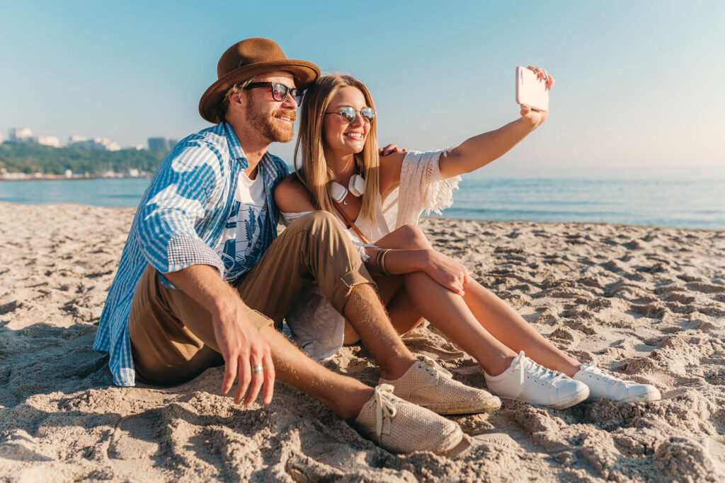 visit sithonia greece young-attractive-smiling-happy-man-woman-sunglasses-sitting-sand-beach-taking-selfie-photo